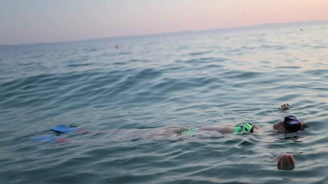 Woman wearing diving mask and blue fins floats calmly on her back in the sea enjoying peaceful sunset water while relaxing after snorkeling and watching the evening skies
