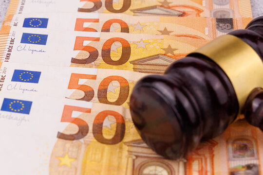 Direct view of a dark mahogany judge gavel resting on structured fifty Euro cash bills over a gray concrete surface, financial corruption investigation and legal fine presentation