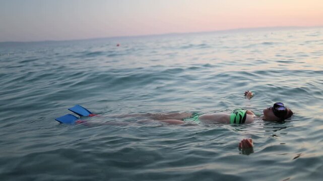 Woman wearing diving mask and blue fins floats peacefully on her back in calm sea at sunset resting after snorkeling and enjoying a serene relaxing evening swim outdoorss