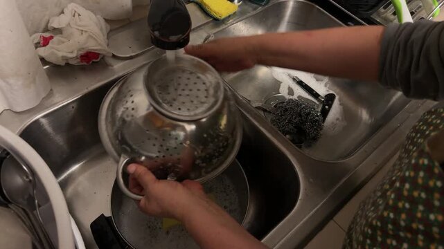 A woman washes used dishes by hand in a kitchen sink while cleaning plates and cookware after food service