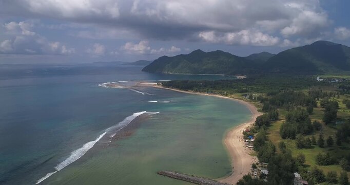 Crescent bay with shallow coral reef pale sand and rainforest mountains as a high drone glides along the Banda Aceh coast revealing inland ponds scattered homes and layered monsoon clouds