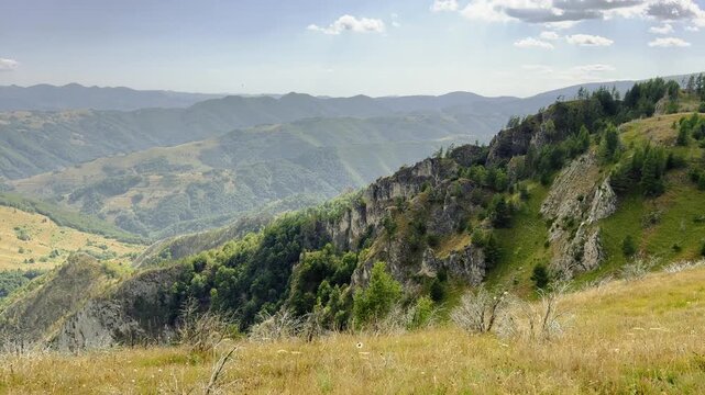 Spectacular wide shot from a high-altitude mountain plateau, showing rugged rocky slopes, lush green forests, and rolling mountain ranges stretching into the horizon under a cloudy sky