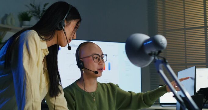 Two female IT support agents working on keyboards at a desk. Asian colleagues communicating through microphones, providing technical assistance and answering client questions.