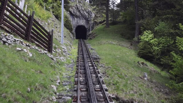 View of the steep cogwheel railway tracks leading into a dark mountain tunnel on Mount Pilatus, surrounded by lush green slopes and pine trees in Alpnach, Obwalden, Switzerland.