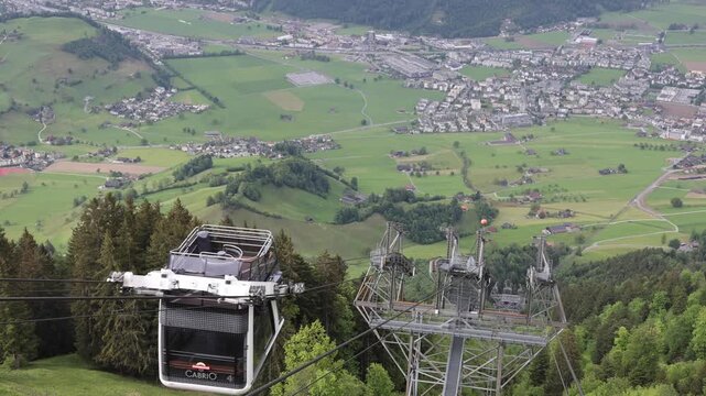 View of the Stanserhorn-Bahn cable car ascending the mountain with lush green alpine meadows and the town of Stans in the background in Stans, Nidwalden, Switzerland.