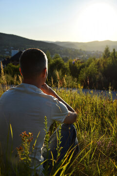 Lonely man sitting outdoors in grass contemplating and looking at sunset in mountains