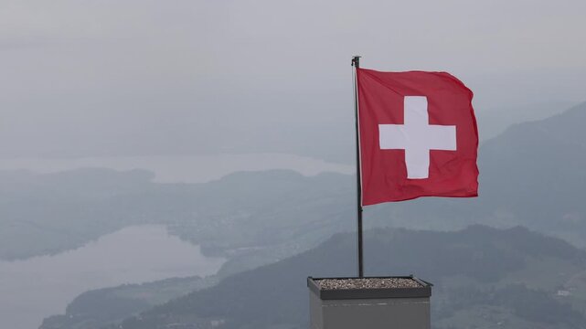 View of Swiss flag waving on a pole at the summit of Stanserhorn with a view of the lake and mountains in the background in Stans, Nidwalden, Switzerland.