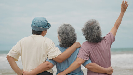 Fototapeta premium Three senior Asian women stand side by side from behind on a beach, embracing each other's waists while one woman enthusiastically waves her hand in the air