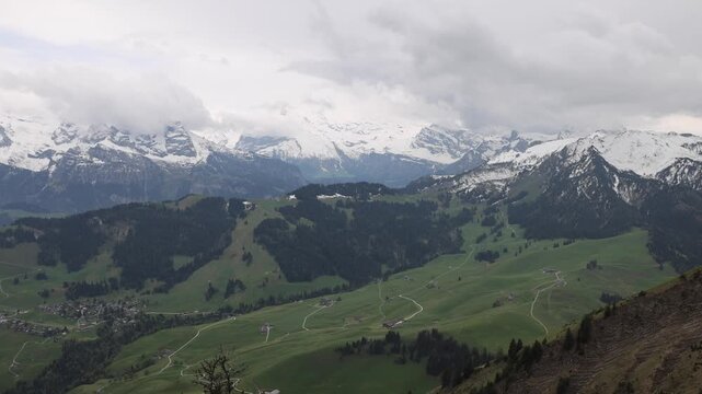 View of Stanserhorn-Bahn mountain landscape with snow-capped peaks, lush green alpine meadows, and winding paths under a cloudy sky in Stans, Nidwalden, Switzerland.