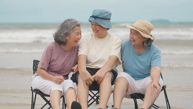 Three elderly Asian women sit together on folding chairs at the beach, laughing happily with ocean waves crashing behind them under an overcast sky