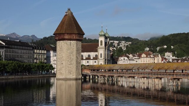 View of the historic Chapel Bridge and Wasserturm spanning the Reuss River in Lucerne, with the Jesuit Church in the background under a clear blue sky in Lucerne, Lucerne, Switzerland.