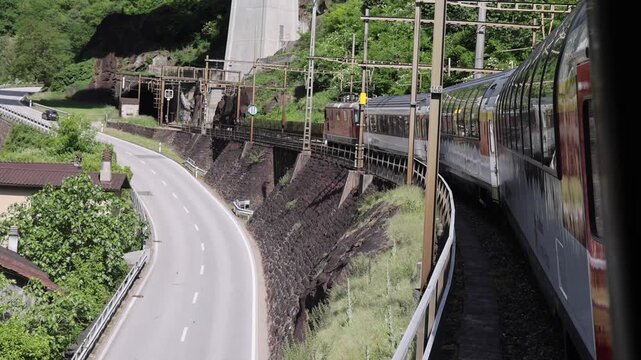 View of the Gotthard Panorama Express SBB train traveling along the tracks with overhead lines and concrete supports above a road in Lugano, Ticino, Switzerland.