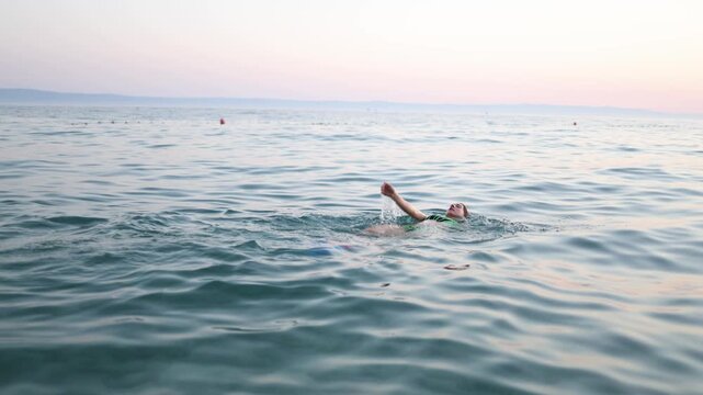 Young woman swims on her back in calm sea water at sunset raising one arm above waves while enjoying freedom peaceful summer vacation and relaxing coastal scenery at dusk