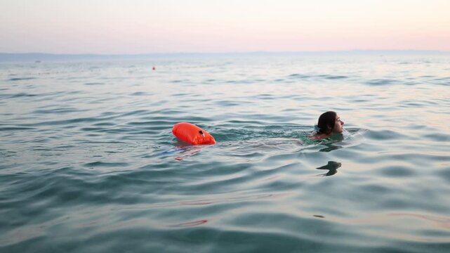 Young woman swims in open sea at sunset towing a bright safety buoy behind her for visibility and protection while enjoying active summer vacation in calm coastal water