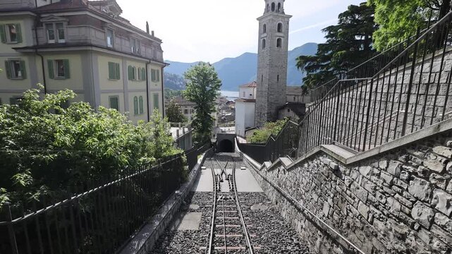 View of the funicular railway tracks leading into a tunnel with the historic San Lorenzo Cathedral bell tower and Lake Lugano in the background in Lugano, Ticino, Switzerland.