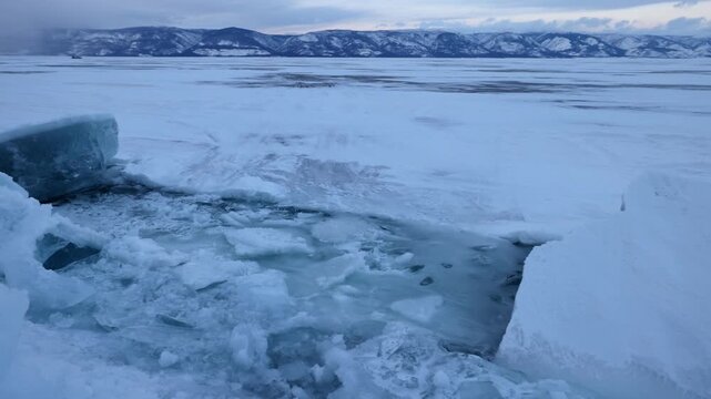 View of the frozen surface of Lake Baikal with jagged ice blocks and snow-covered mountains in the distance in Khuzhir, Irkutsk Oblast, Russia.