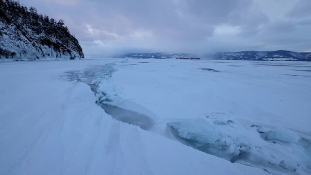 View of the frozen surface of Lake Baikal with snow-covered ice and rocky shores under a cloudy sky in Khuzhir, Irkutsk Oblast, Russia.