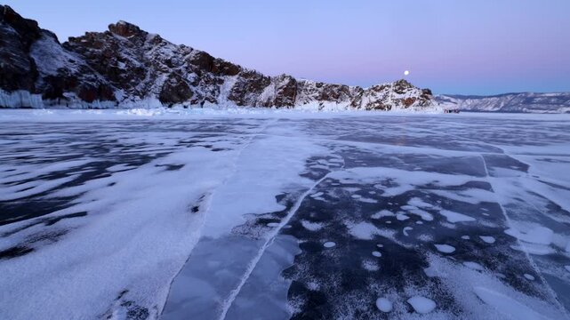View of the frozen surface of Lake Baikal with snow-covered rocky cliffs under a twilight sky in Khuzhir, Irkutsk Oblast, Russia.