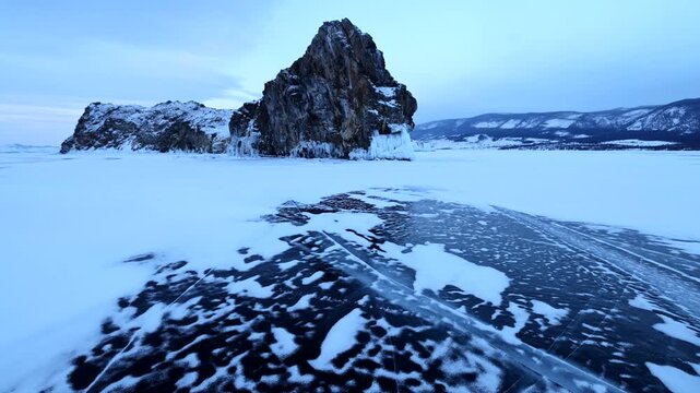 View of the iconic Shamanka Rock on the frozen surface of Lake Baikal with cracked ice and snow-covered landscape in Khuzhir, Irkutsk Oblast, Russia.
