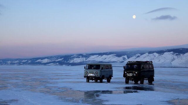 View of two UAZ vans parked on the frozen surface of Lake Baikal with snowy mountains in the background under a pale moon in Khuzhir, Irkutsk Oblast, Russia.