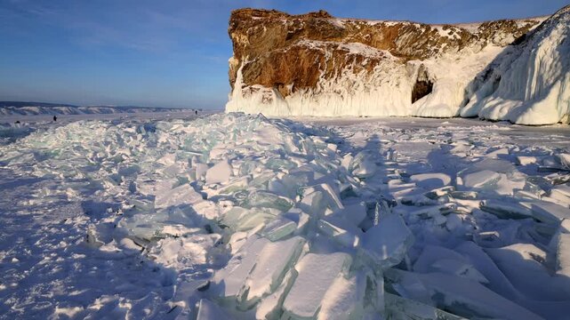 View of the frozen surface of Lake Baikal with jagged ice shards and snow-covered cliffs in the background in Khuzhir, Irkutsk Oblast, Russia.