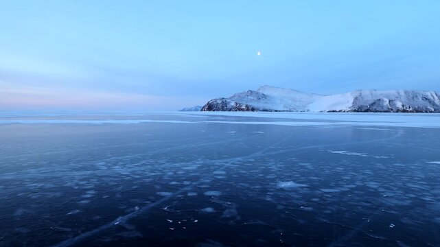 View of the frozen surface of Lake Baikal with snow-covered hills under a twilight sky in Khuzhir, Irkutsk Oblast, Russia.