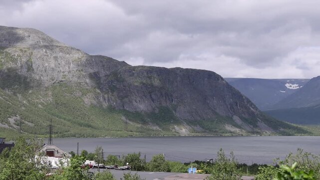 View of the Khibiny Mountains rising above a lake with lush green vegetation and a cloudy sky in Kirovsk, Murmansk Oblast, Russia.