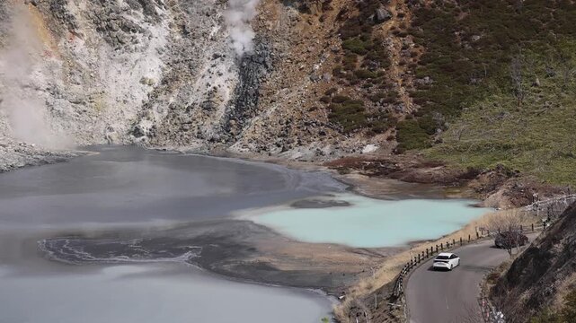 View of Noboribetsu Jigokudani Valley with steaming volcanic vents and turquoise geothermal pools along a winding road in Noboribetsu, Hokkaido, Japan.