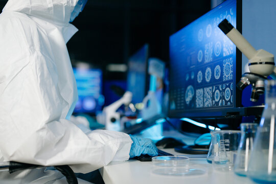 Medical researcher in protective suit reviewing and recording data from multi screen digital displays inside research laboratory.