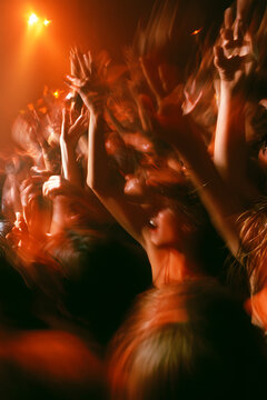 Dancers in a crowded nightclub raise their hands beneath warm orange lights, their movements blurred close-up with a lively effect. Motion blur, selective focus.