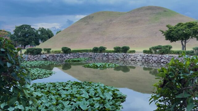 View of the historic Daereungwon Tomb Complex featuring a large grassy burial mound reflecting in a tranquil pond with lily pads and stone walls in Gyeongju, Gyeongsangbuk-do, South Korea.