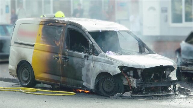 Vehicle fire aftermath with burned van, smoke and small flames beneath chassis. Destroyed commercial vehicle on urban street with fire hoses across wet asphalt. Charred van with severe heat damage