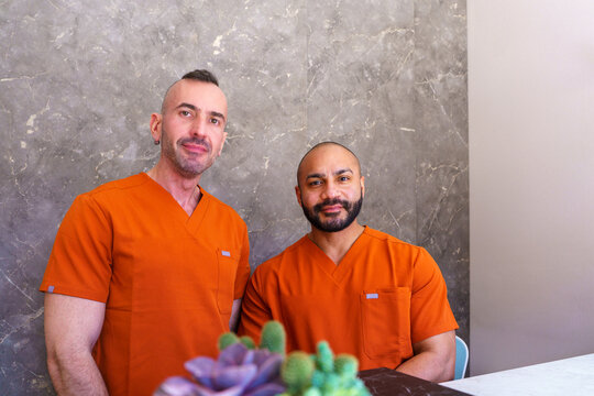 Male beauticians in orange scrubs standing at reception desk of modern beauty clinic, smiling at camera