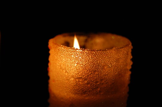 Close-up of a burning candle with a bright glowing flame reflected in a dark surface, isolated against a black background. Atmospheric low-key lighting creates a warm, dramatic and mysterious mood.