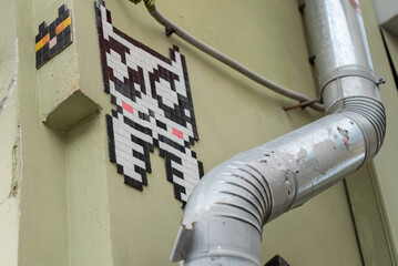 A pixel art cat mural on a green wall, next to a metal drainpipe. The cat features a black and white design with pink accents. © Ksu