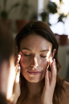 A young woman with freckled skin rests her fingertips on her cheeks with eyes closed, surrounded by houseplants in warm indoor light during a calm self care moment.