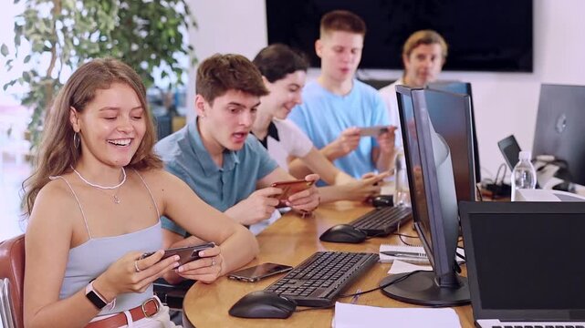 Cheerful young female student studying together with coursemates in computer workshop, actively using mobile phone to access online learning materials or participating in group discussions