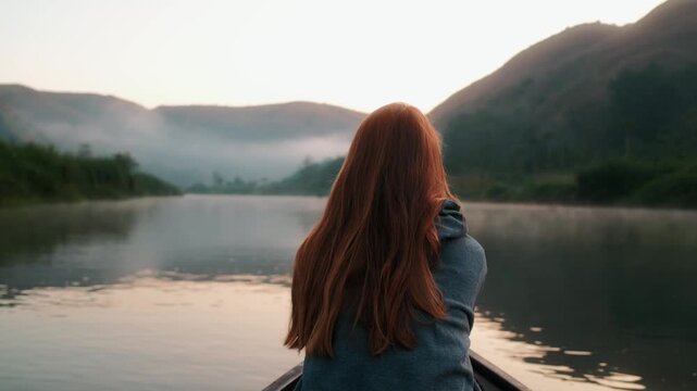 View of a woman with red hair sitting in a boat on a calm river surrounded by misty hills at sunrise in Kecamatan Kambera, Nusa Tenggara Timur, Indonesia.