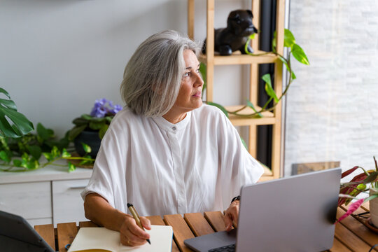 Mature white woman writing notes with laptop in home office in Spain