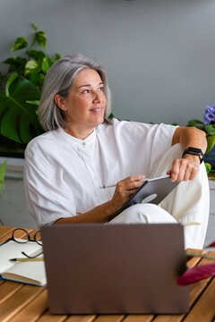 Mature white woman using digital tablet in home workspace with plants