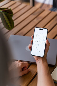 Hand holding smartphone with digital calendar app and laptop on wooden