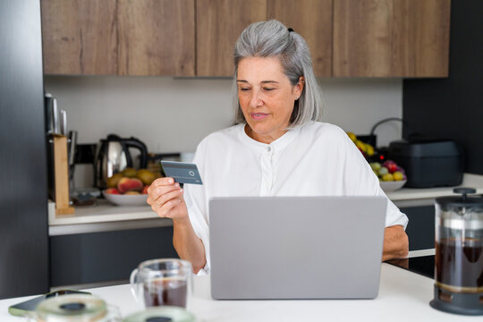 Senior white woman making online payment on laptop in kitchen in Spain
