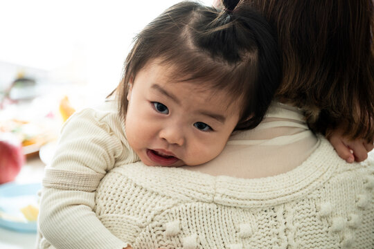 Cute Asian Toddler Girl Hugging Mother's Shoulder Looking at Camera Indoors