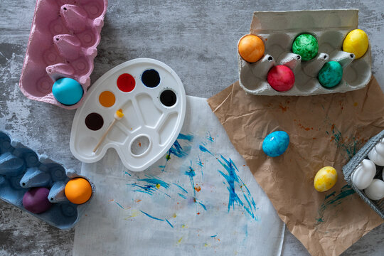 Overhead Easter craft composition featuring colorful decorative eggs a watercolor palette paintbrushes and additional craft materials arranged on a rustic blue and gray background