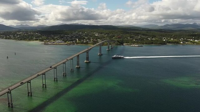 Aerial drone footage of the Gisund Bridge crossing the Gisundet strait between Finnsnes and Senja in Norway, with a ferry passing beneath and a coastal town in the background