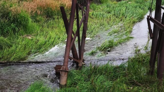 A shallow stream flows steadily through a grassy area, flanked by rusted metal structures. Gentle currents create ripples and foam over rocks, while lush green vegetation lines the banks.