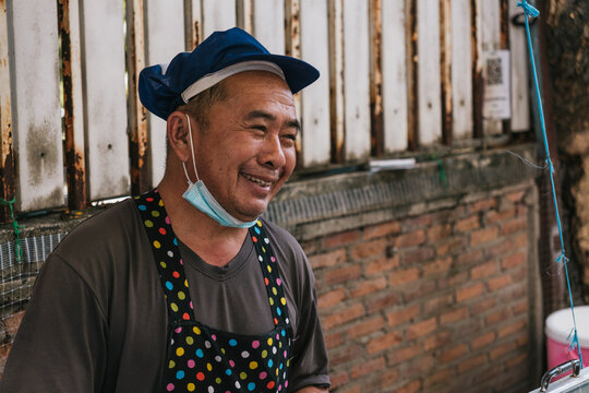 Smiling Asian street food vendor wearing a blue chef hat and colorful polka dot apron with face mask pulled down