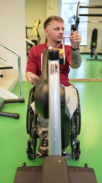 Disabled man in wheelchair doing exercise for upper body strength on adaptive sports equipment at gym, personalized training session for people with spinal injuries, male using ergometer machine.
