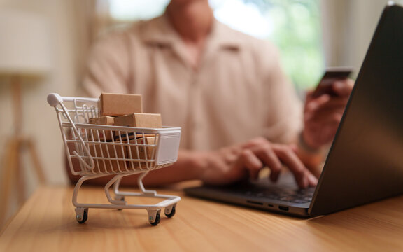 Asian Man shops online with a laptop and payment card beside a miniature cart filled with parcels, representing e-commerce, digital payment, delivery, checkout, online orders and consumer shopping.