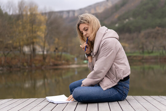 Thoughtful woman writing notes on paper outside by calm lake